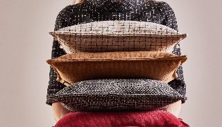 Camira Monochrome Sett kumaşıyla kaplı ızgara ve dokuma desenli yastıklar, taba ve kırmızı düz minderlerle bir kişinin kucağında