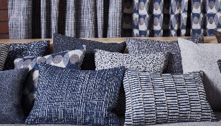 Camira Patternmaker yün kumaşının ızgara, nokta, çizgi ve daire desenli yastıkları ve perdeleri, mavi ve gri tonlarında tuğla duvar önünde