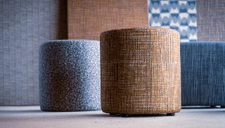 Camira Patternmaker kumaşıyla döşenmiş hardal ve gri desenli yuvarlak tabureler, arkada ızgara ve geometrik desenli paneller