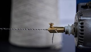 Camira Main Line Twist'in bükümlü ipliğinin pirinç büküm aparatından geçtiği yakın çekim, arkada iplik konisi