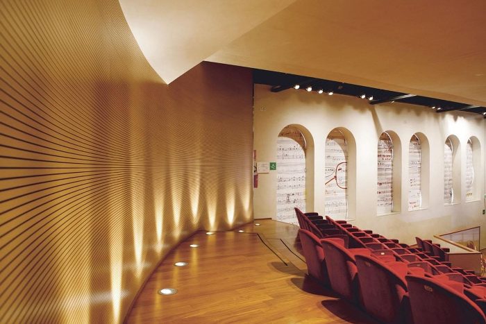 Konser salonunda kavisli açık ahşap yatay derzli akustik duvar, kırmızı koltuklar ve nota vitrinleri