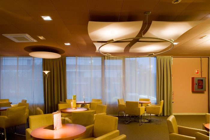Otel lobisinde yelken biçimli beyaz akustik kanopi paneli ve daire disk, sarı koltuklu oturma alanı