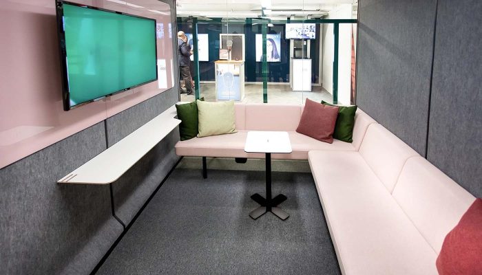 Akustik toplantı kabini iç düzeni, pembe L koltuk, duvar rafı ve ekran ile showroom uygulaması