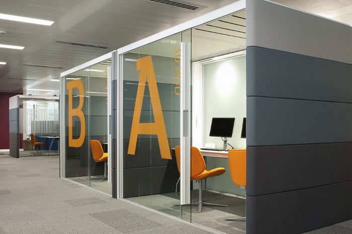 Bağımsız ofis bölümü cam cephesinde büyük turuncu harf grafikleri bulunan kabin sırası, içlerinde monitörlü masalar ve turuncu sandalyeler