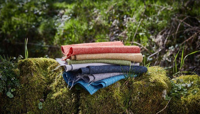 Camira Hemp yün ve kenevir kumaşının mercan, bej, yeşil, gri ve mavi tonlardaki örnekleri, doğada yosunlu taş üzerinde yığın halinde