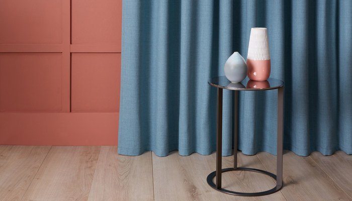 Camira Sumi mavi yün kumaşından zemine uzanan perde, terracotta panelli duvar önünde metal yan masa ve vazolarla