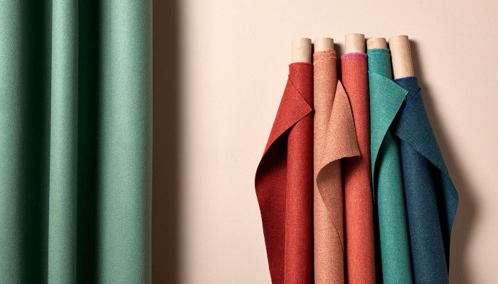 Camira Main Line Flax kumaşının kırmızı, mercan, petrol ve mavi tonlardaki ruloları, açık pembe duvar önünde