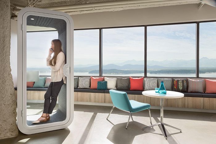 Telefon görüşme kabini beyaz gövdesiyle panoramik manzaralı ofis katında, içeride ayakta telefonla konuşan bir kullanıcı