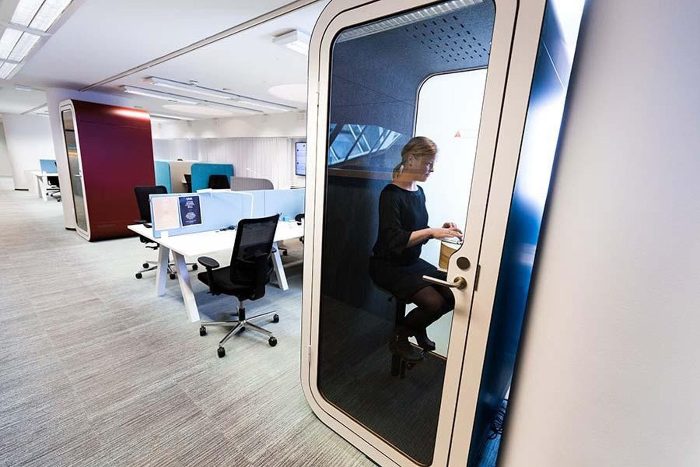 Ofis telefon kabini krem kapı çerçevesiyle açık ofiste, içeride laptopla çalışan kullanıcı, arka planda bordo modül