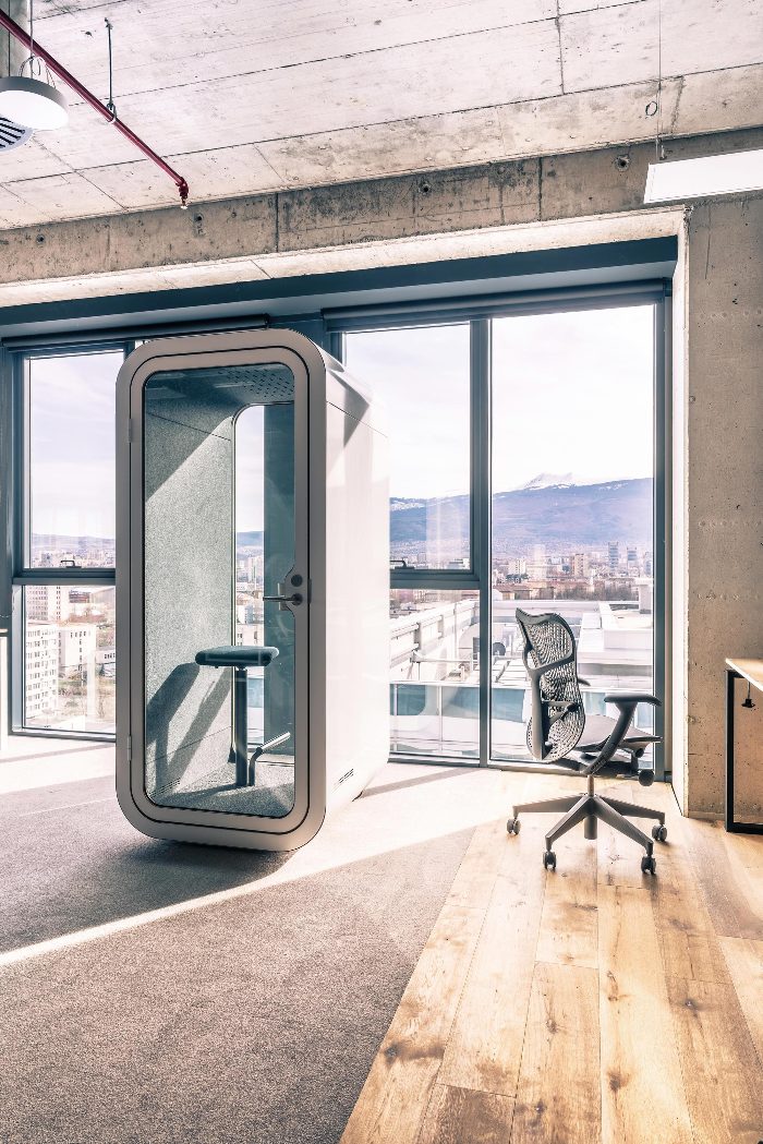 Telefon görüşme kabini açık gri gövdesiyle beton tavanlı loft ofiste, şehir manzaralı cam cephe önünde perspektif görünüm